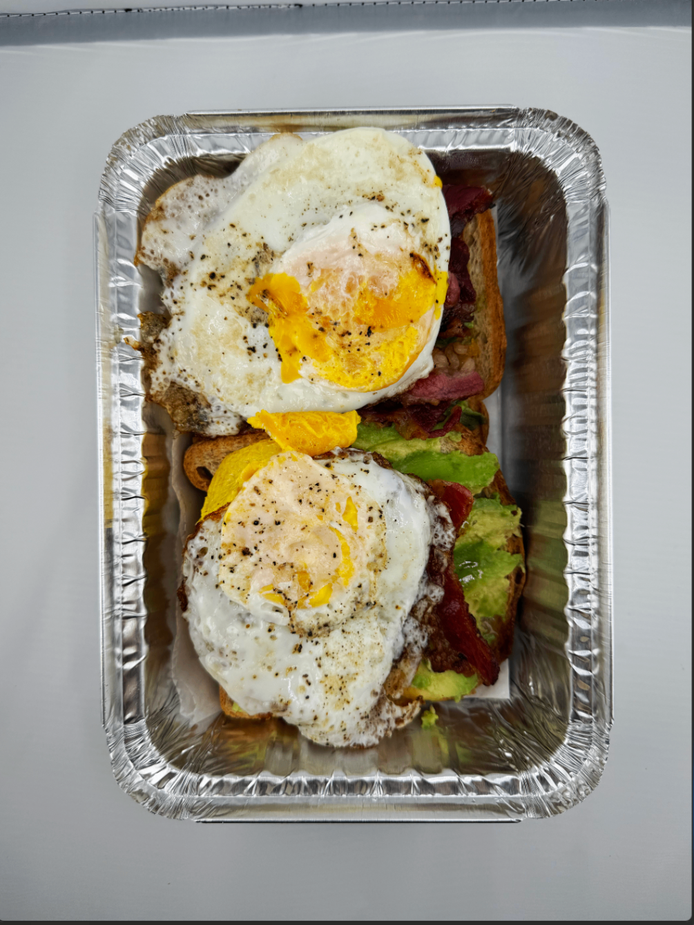 Avocado toast topped with beef bacon and two fried eggs in a takeout tray